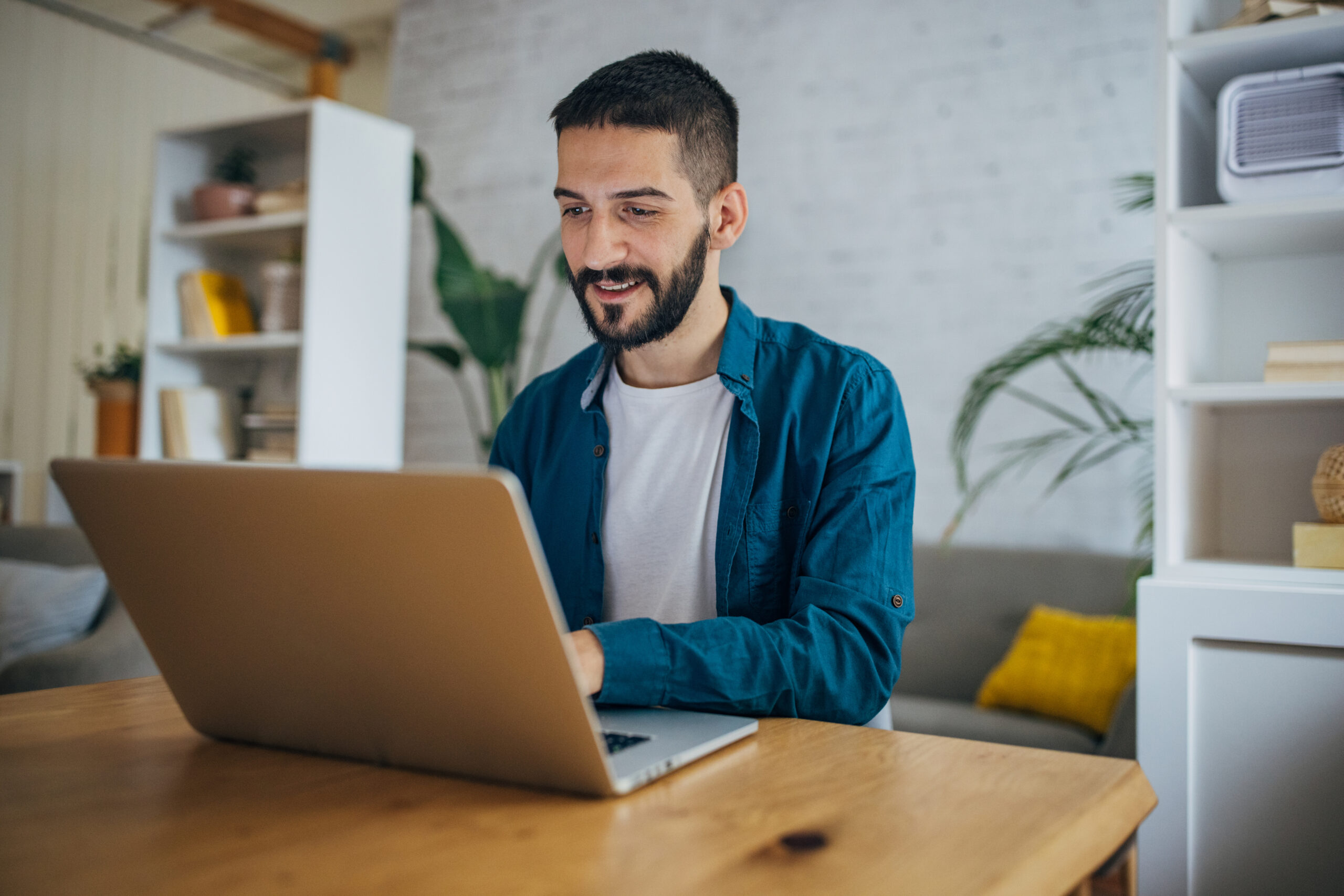 A man wearing a blue shirt types on his laptop while sitting at a wooden desk in his home office.